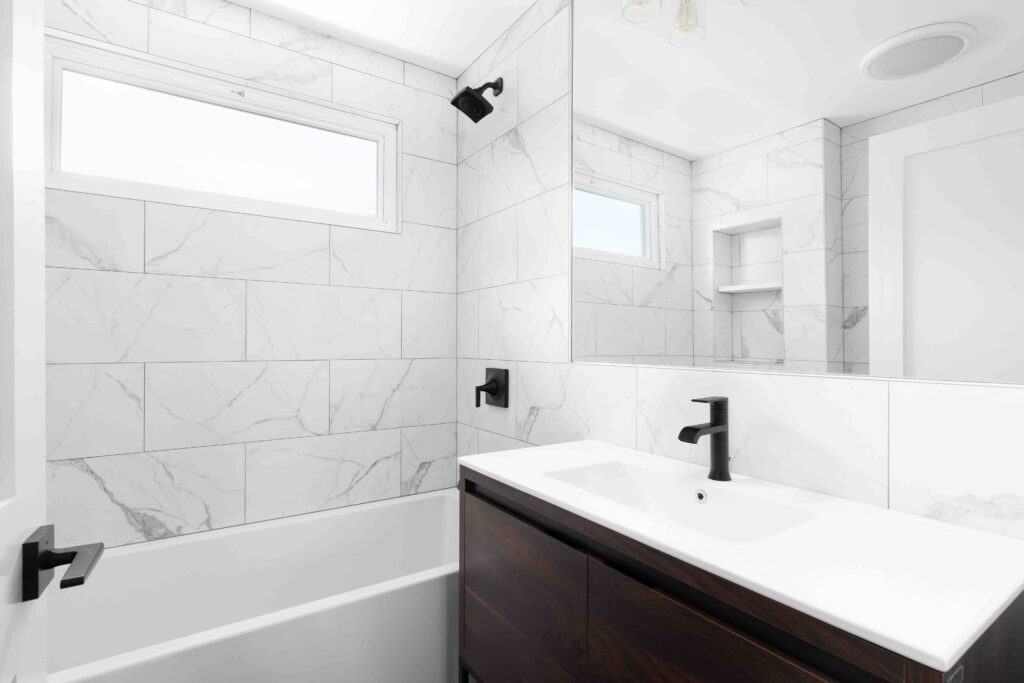 A renovated bathroom with a wood vanity cabinet, white marble sink and large marble tiles lining the shower walls.