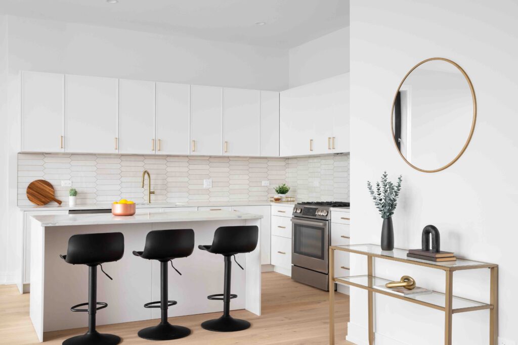 A kitchen detail with white cabinets, a picket tile backplash, white bar stools sitting at the island, and decorations under a circular mirror. No brands or labels.