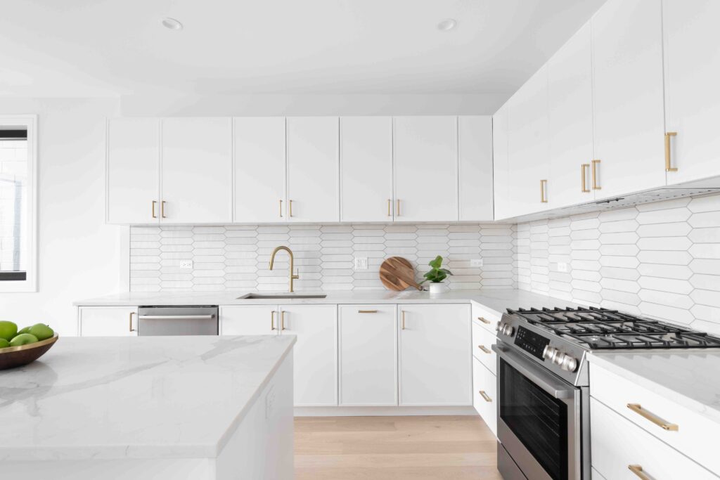 A beautiful kitchen detail with white cabinets, a gold faucet, white marble countertops, and a brown picket ceramic tile backsplash.