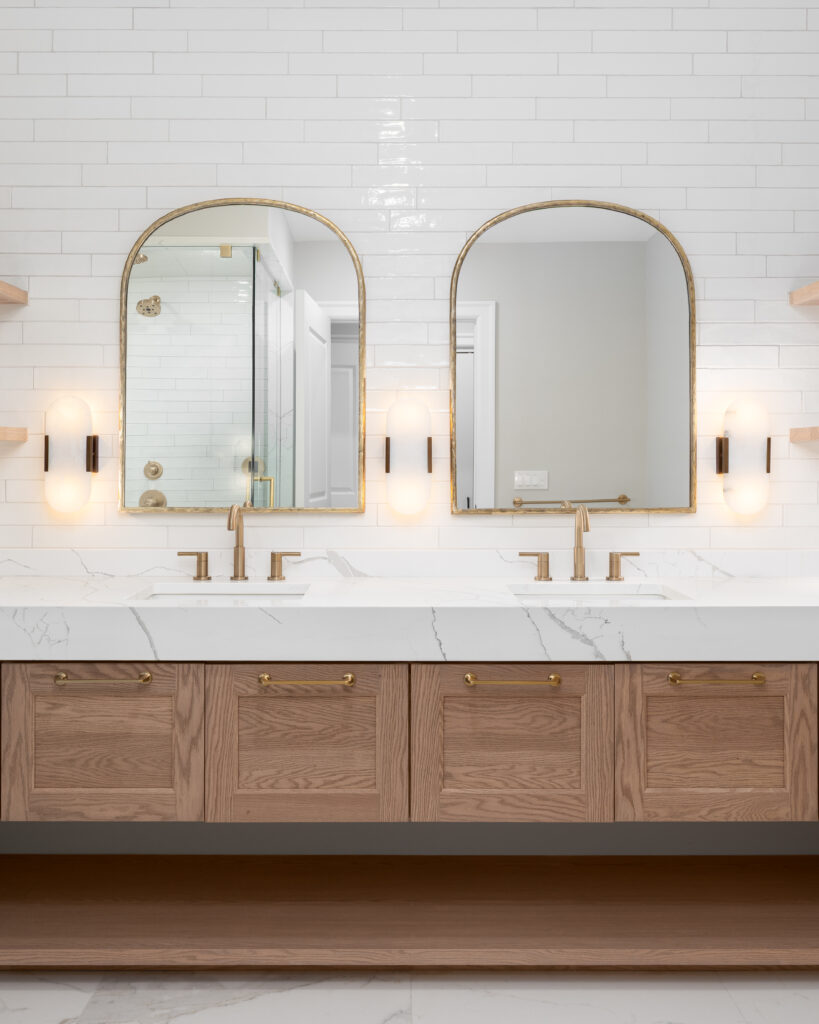 A bathroom detail with gold faucets, light fixtures, and mirrors, a wood cabinet, large marble slab countertop, and a subway tile wall.